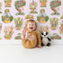 Lovely child standing in front of Whimsy Tots Curious Critter Village Wallpaper in Blush Pink with house, cat, tree, cup, jar, ladder, tower in design.