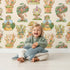 Lovely child standing in front of Whimsy Tots Curious Critter Village Wallpaper in Beige with house, cat, cup, tree, jar, tower in design.