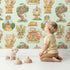 Lovely child standing in front of Whimsy Tots Curious Critter Village Wallpaper in Green with house, cat, tree, cup, ladder, jar, tower in design.