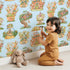 Lovely child standing in front of Whimsy Tots Curious Critter Village Wallpaper in Blue with house, cat, tree, cup, jar, ladder, tower in design.