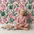 Lovely child standing in front of Whimsy Tots Enchanted Meadow Prance Wallpaper in Beige with unicorn, flower, tulip, leaf in design.