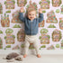Lovely child standing in front of Whimsy Tots Secret Garden Cutaway Homes Wallpaper in Blush Pink with house, mushroom, vegetable, root, tree in design.