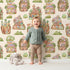 Lovely child standing in front of Whimsy Tots Secret Garden Cutaway Homes Wallpaper in Beige with house, mushroom, tree, gourd, vegetable in design.