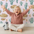 Lovely child standing in front of Whimsy Tots Cactus Cloud Voyage Wallpaper in Blush Pink with cactus, airship, balloon, moon in design.