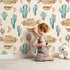 Lovely child standing in front of Whimsy Tots Cactus Cloud Voyage Wallpaper in Sand with cactus, airship, balloon, moon in design.