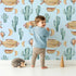 Lovely child standing in front of Whimsy Tots Cactus Cloud Voyage Wallpaper in Sky Blue with cactus, airship, balloon, moon in design.