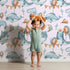 Lovely child standing in front of Whimsy Tots Ocean Pals Celebration Wallpaper in Blush Pink with whale, balloon, octopus, coral, bunting in design.