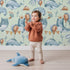 Lovely child standing in front of Whimsy Tots Ocean Pals Celebration Wallpaper in Mint Green with whale, balloon, octopus, coral, bunting in design.
