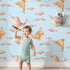 Lovely child standing in front of Whimsy Tots Blushing Ocean Friends Wallpaper in Blue with whale, dolphin, narwhal, fish, wave in design.