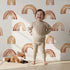 Lovely child standing in front of Whimsy Tots Boho Brush Rainbow Wallpaper in White with rainbow, stripe, arch in design.