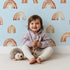 Lovely child standing in front of Whimsy Tots Boho Brush Rainbow Wallpaper in Blue with rainbow, stripe, arch in design.