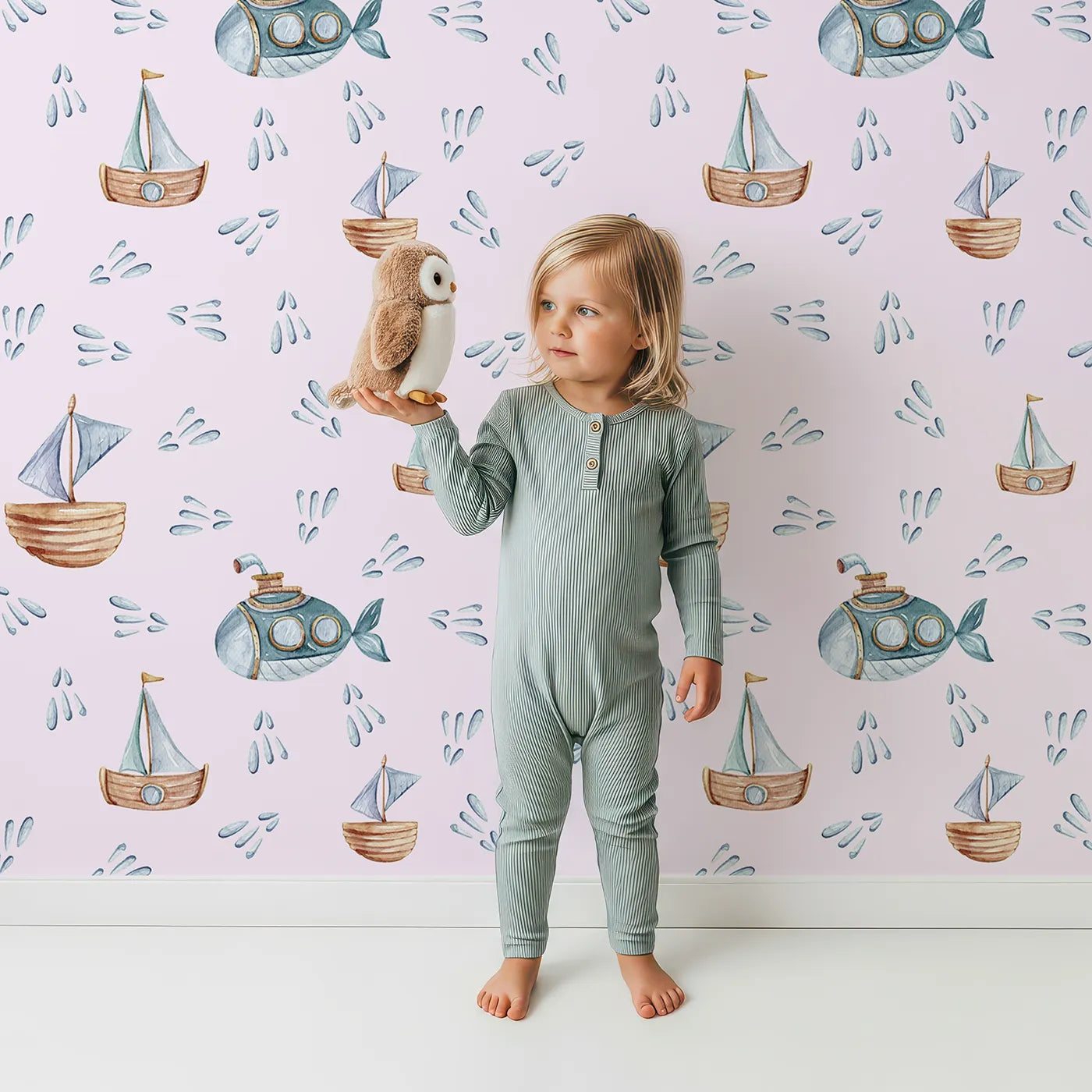 Lovely child standing in front of Whimsy Tots Seaside Vessel Parade Wallpaper in Pink with boat, submarine, splash, sail in design.