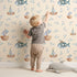 Lovely child standing in front of Whimsy Tots Seaside Vessel Parade Wallpaper in Beige with boat, submarine, splash, sail in design.