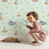 Lovely child standing in front of Whimsy Tots Seaside Vessel Parade Wallpaper in Mint Green with boat, submarine, splash, sail in design.