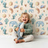Lovely child standing in front of Whimsy Tots Balloon Friends Parade Wallpaper in Beige with dinosaur, elephant, turtle, cactus, moon, heart, rainbow in design.