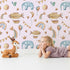 Lovely child standing in front of Whimsy Tots Sky Dreams Celebration Wallpaper in Pink with airship, balloon, crown, moon, rainbow, heart, elephant, bird in design.