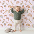 Lovely child standing in front of Whimsy Tots Sky Bloom Party Wallpaper in Pink with rainbow, balloon, moon, star, bunting, heart in design.