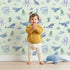 Lovely child standing in front of Whimsy Tots Whale Song Friends Wallpaper in Mint Green with whale, fish, narwhal, octopus, jellyfish in design.