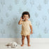 Lovely child standing in front of Whimsy Tots Petal Outline Grace Wallpaper in Blue with flower, leaf, sprig, botanical in design.
