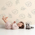 Lovely child standing in front of Whimsy Tots Petal Bloom Grace Wallpaper in Sand with flower, leaf, blossom, sprig in design.