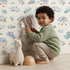 Lovely child standing in front of Whimsy Tots Cloudy Dreams Parade Wallpaper in White with cloud, star, sheep, sky in design.