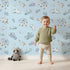 Lovely child standing in front of Whimsy Tots Cloudy Dreams Parade Wallpaper in Blue with cloud, star, sheep, sky in design.