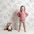 Lovely child standing in front of Whimsy Tots Petal Outline Bloom Wallpaper in White with flower, petal, bell flower, sprig, leaf, botanical in design.