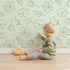 Lovely child standing in front of Whimsy Tots Petal Outline Bloom Wallpaper in Green with flower, petal, bell flower, sprig, leaf, botanical in design.