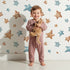 Lovely child standing in front of Whimsy Tots Starlight Scribbles Wallpaper in White with star, scribble, doodle in design.