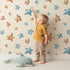 Lovely child standing in front of Whimsy Tots Starlight Scribbles Wallpaper in Sand with star, scribble, doodle in design.