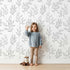 Lovely child standing in front of Whimsy Tots Wild Leafline Garden Wallpaper in White with leaf, botanical, sprig, berry, line art in design.