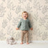 Lovely child standing in front of Whimsy Tots Wild Leafline Garden Wallpaper in Sand with leaf, botanical, sprig, berry, line art in design.