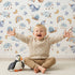 Lovely child standing in front of Whimsy Tots Dreamscape Journey Wallpaper in White with narwhal, snail, whale, sheep, moon, star, rainbow, boat, cloud in design.