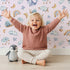 Lovely child standing in front of Whimsy Tots Dreamscape Journey Wallpaper in Blush Pink with narwhal, snail, whale, sheep, moon, star, rainbow, boat, cloud in design.