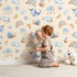 Lovely child standing in front of Whimsy Tots Dreamscape Journey Wallpaper in Sand with narwhal, snail, whale, sheep, moon, star, rainbow, boat, cloud in design.