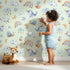 Lovely child standing in front of Whimsy Tots Dreamscape Journey Wallpaper in Mint Green with narwhal, snail, whale, sheep, moon, star, rainbow, boat, cloud in design.