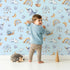 Lovely child standing in front of Whimsy Tots Dreamscape Journey Wallpaper in Blue with narwhal, snail, whale, sheep, moon, star, rainbow, boat, cloud in design.