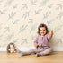 Lovely child standing in front of Whimsy Tots Leaf Outline Symphony Wallpaper in Beige with leaf, branch, botanical, outline in design.