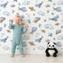 Lovely child standing in front of Whimsy Tots Sleepy Sea Sky Wallpaper in White with whale, turtle, fish, cloud, star, heart in design.