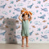 Lovely child standing in front of Whimsy Tots Sleepy Sea Sky Wallpaper in Blush Pink with whale, turtle, fish, cloud, star, heart in design.