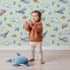 Lovely child standing in front of Whimsy Tots Sleepy Sea Sky Wallpaper in Mint Green with whale, turtle, fish, cloud, star, heart in design.