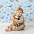 Lovely child standing in front of Whimsy Tots Sleepy Sea Sky Wallpaper in Blue with whale, turtle, fish, cloud, star, heart in design.