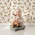 Lovely child standing in front of Whimsy Tots Botanical Numbers Wallpaper in Sand with number, leaf, vine, foliage in design.