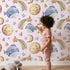 Lovely child standing in front of Whimsy Tots Sleepy Celestial Whale Wallpaper in Blush Pink with whale, moon, star, fish, comet in design.