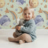 Lovely child standing in front of Whimsy Tots Sleepy Celestial Whale Wallpaper in Mint Green with whale, moon, star, fish, comet in design.