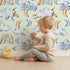 Lovely child standing in front of Whimsy Tots Sleepy Cloud Parade Wallpaper in Mint Green with cloud, moon, sheep, rainbow, star in design.