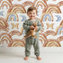 Lovely child standing in front of Whimsy Tots Boho Rainbow Night Wallpaper in White with rainbow, moon, heart, arch in design.