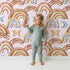 Lovely child standing in front of Whimsy Tots Boho Rainbow Night Wallpaper in Blush Pink with rainbow, moon, heart, arch in design.