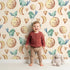 Lovely child standing in front of Whimsy Tots Lunar Lullaby Birds Wallpaper in Sand with bird, moon, heart in design.