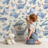 Lovely child standing in front of Whimsy Tots Sleepy Sea Friends Wallpaper in Sand with whale, narwhal, fish in design.